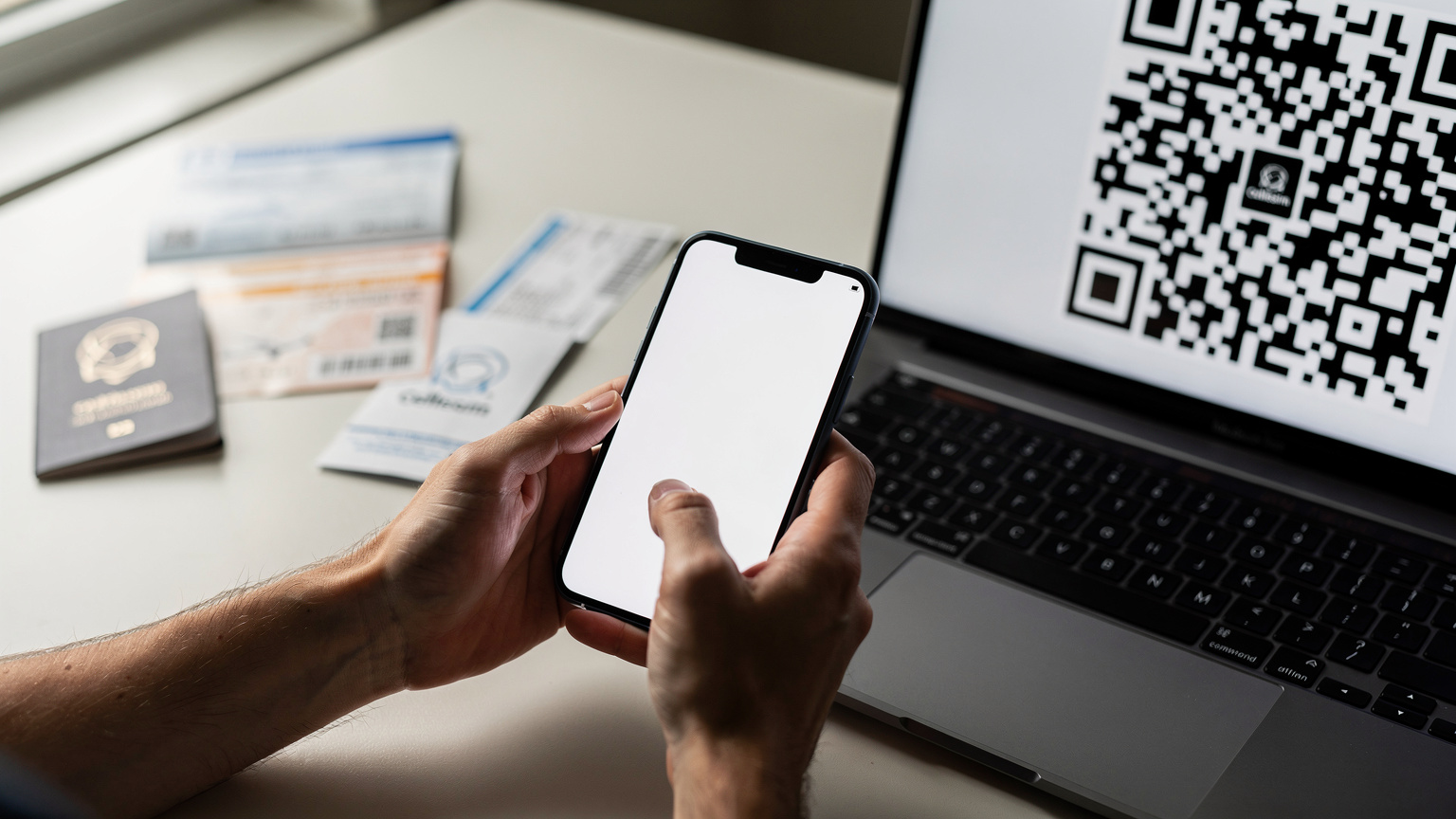 An adult's hands holding a smartphone, scanning a QR code displayed on a computer screen, indicating the eSIM activation process, with travel documents slightly visible in the background.