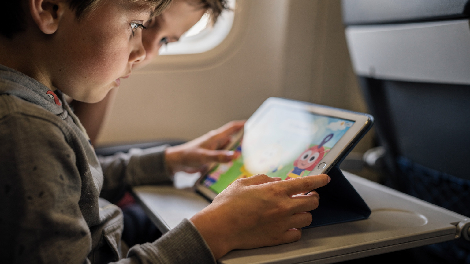 Close-up of a child's hands holding an iPad, with another child peeking over their shoulder, watching a cartoon, possibly on an airplane tray table or in a car.