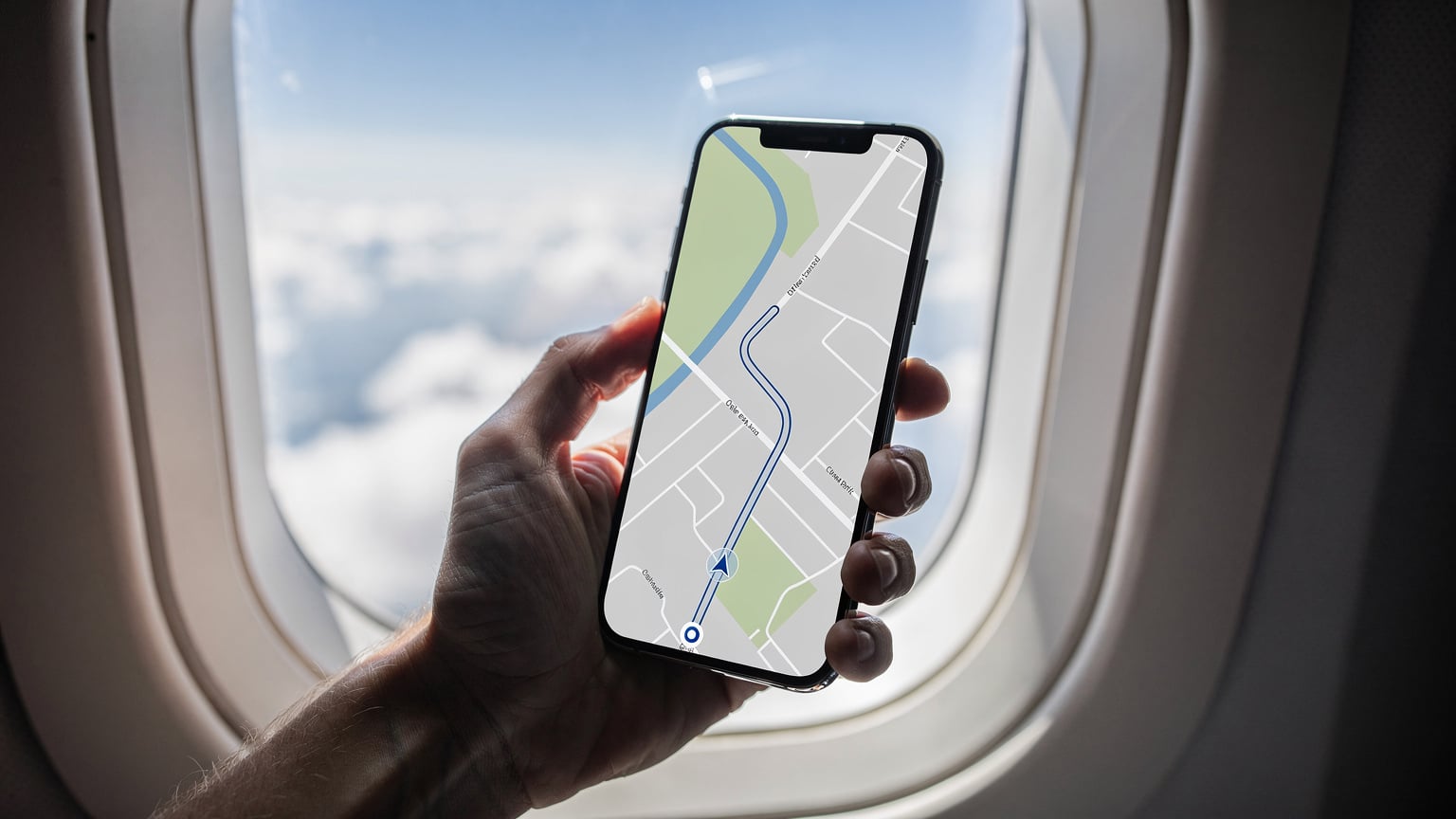 Uma mão segurando um smartphone moderno em frente a uma janela de avião, com a tela mostrando claramente um aplicativo de mapas offline com uma rota de viagem. A paisagem de nuvens é visível ao fundo.