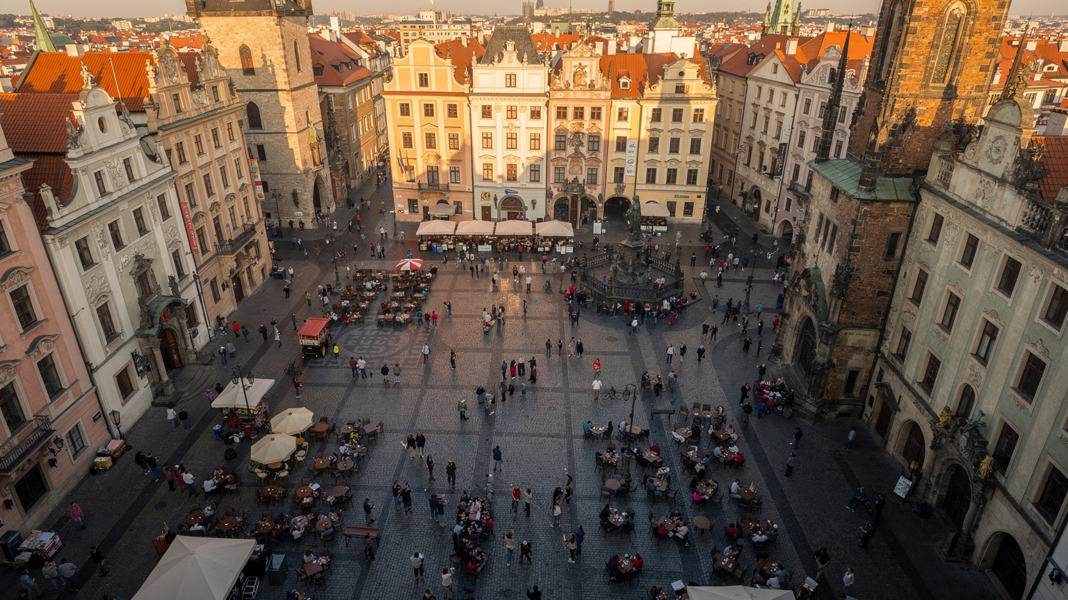 Una veduta aerea di una vivace piazza storica a Praga, Repubblica Ceca, con persone che passeggiano e interagiscono con i loro telefoni, mentre il sole del tardo pomeriggio illumina gli antichi edifici barocchi.
