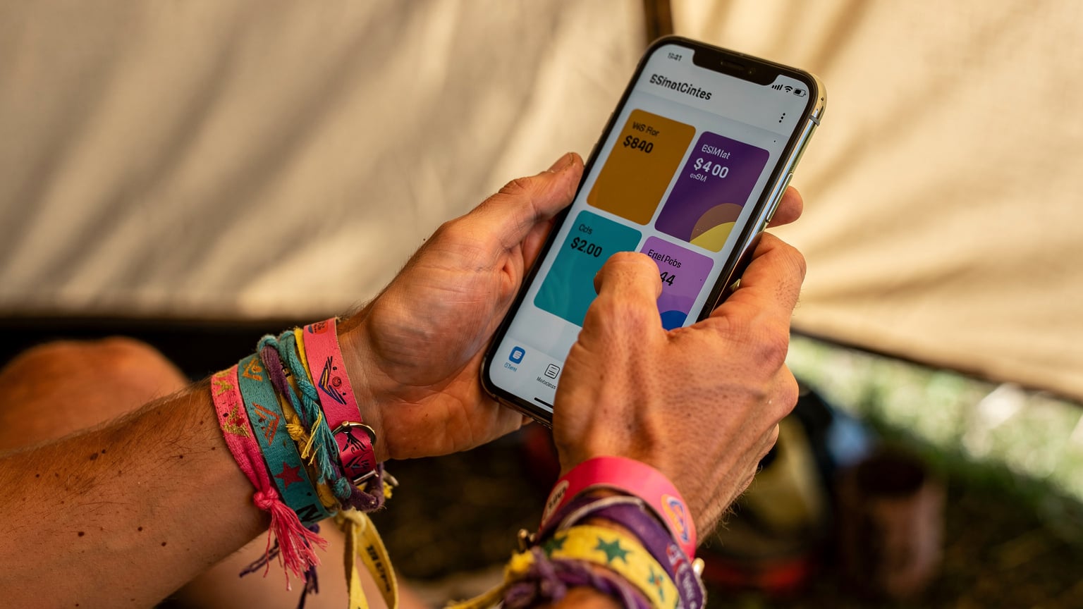 A close-up of a person's hands holding a smartphone, actively comparing eSIM plans on a travel app, with festival wristbands subtly visible on their wrist.