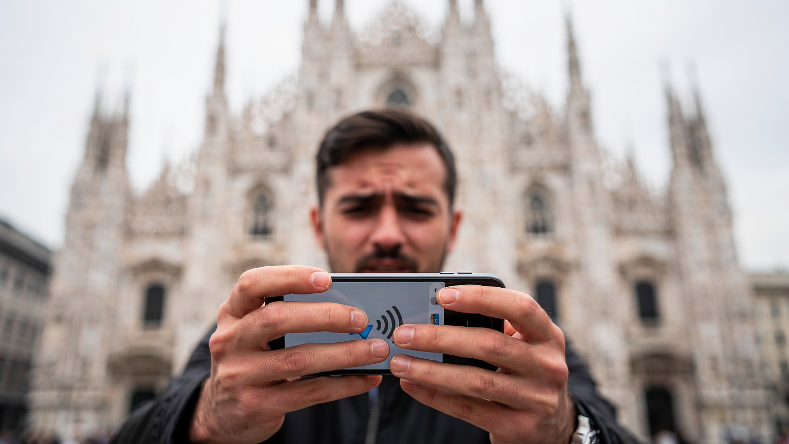 Un primo piano delle mani di una persona che tiene uno smartphone, che mostra una schermata generica di errore di connessione o assenza di segnale. Sullo sfondo, leggermente sfocato, si intravede il maestoso Duomo di Milano, simbolo della città. L'espressione della persona è leggermente frustrata o preoccupata. Scatto con obiettivo 50mm, profondità di campo ridotta per focalizzarsi sulle mani e lo schermo. Fotografia editoriale, stile Wirecutter, che mostra il prodotto in un contesto reale di un problema. Photorealistic, no on-screen text or UI, no readable logos, no superimposed graphics.