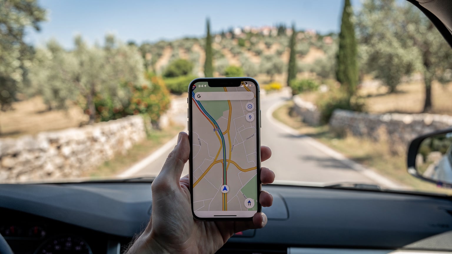 Ένα χέρι κρατά ένα κινητό τηλέφωνο που εμφανίζει έναν χάρτη πλοήγησης, ενώ το αυτοκίνητο κινείται σε έναν γραφικό αγροτικό δρόμο της Κροατίας, με ελαιώνες και μεσογειακή βλάστηση στο βάθος.