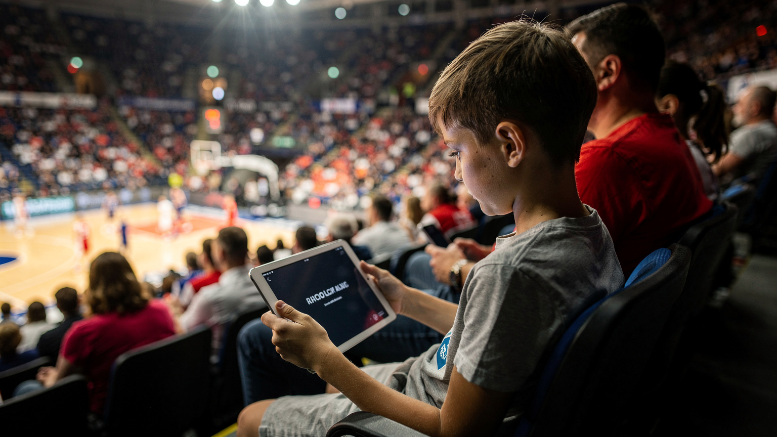 Ένα παιδί 9 ετών με ένα iPad στα χέρια του, παρακολουθεί περιεχόμενο εκτός σύνδεσης, ενώ βρίσκεται σε ένα πολυσύχναστο γήπεδο μπάσκετ, με τους γονείς του να παρακολουθούν τον αγώνα στο παρασκήνιο.