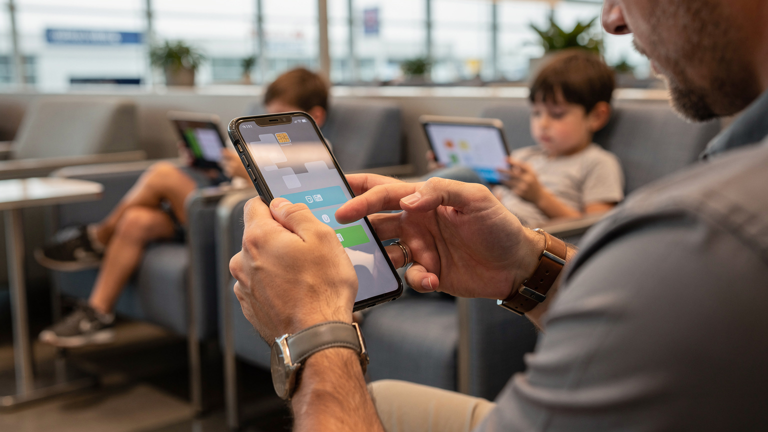 Un padre viajero configurando los ajustes de datos en su smartphone con una eSIM activa, mientras sus hijos juegan tranquilamente en segundo plano, en la sala de espera de un aeropuerto.