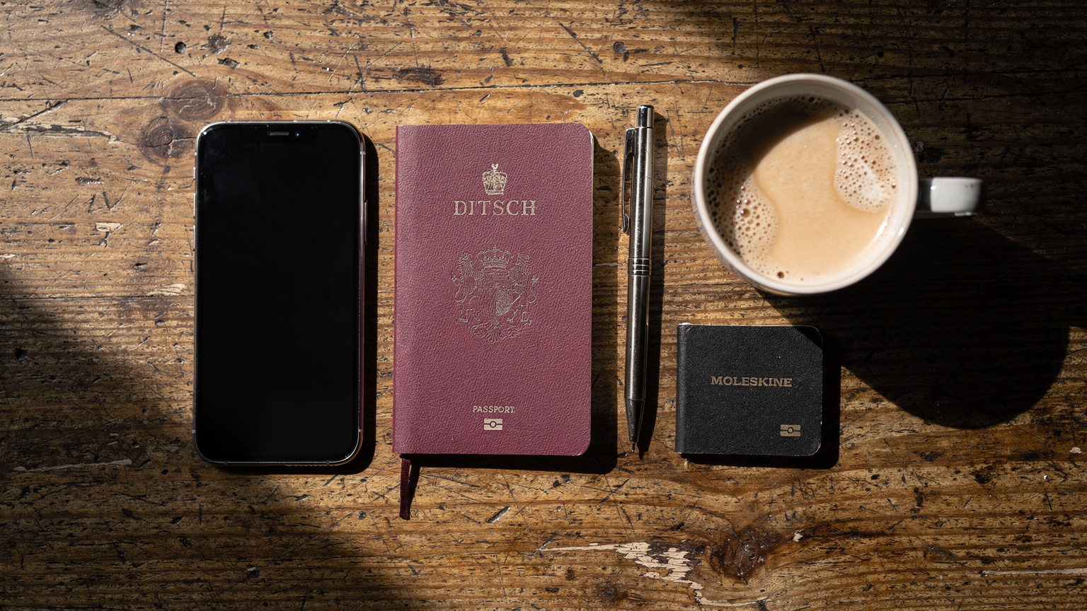 Een georganiseerde flat-lay van een iPhone 18, paspoort, een notitieboekje, pen en een kopje koffie op een houten tafel in een gezellig café in Barcelona, wat een voorbereiding op een reis suggereert.