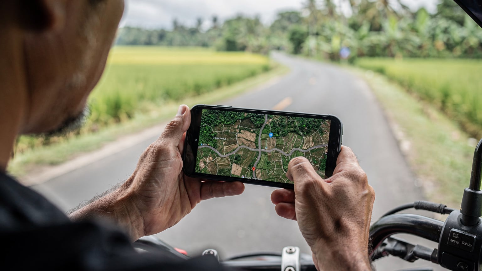 Tangan seorang pria sedang memegang ponsel dengan aplikasi Google Maps dalam mode offline yang menampilkan area Bali, sementara di latar belakang terlihat pemandangan jalan pedesaan yang sepi, mengindikasikan perjalanan di area terpencil.