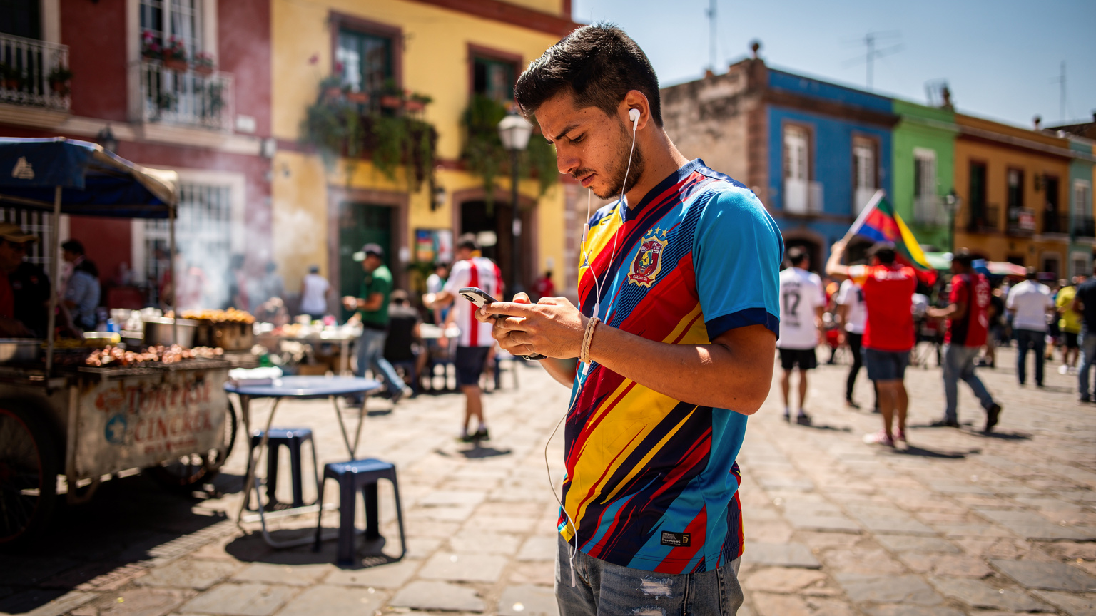 Un aficionado al fútbol latinoamericano usando su teléfono para seguir un partido o buscar direcciones en una bulliciosa plaza de una ciudad anfitriona de la Copa América, con puestos de comida callejera y gente alrededor.
