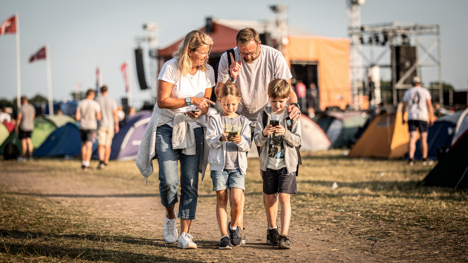 Et par forældre og deres to børn navigerer festivalpladsen ved Roskilde Festival med deres smartphones, mens de tjekker et digitalt kort for at finde vej. Solen skinner på dem.