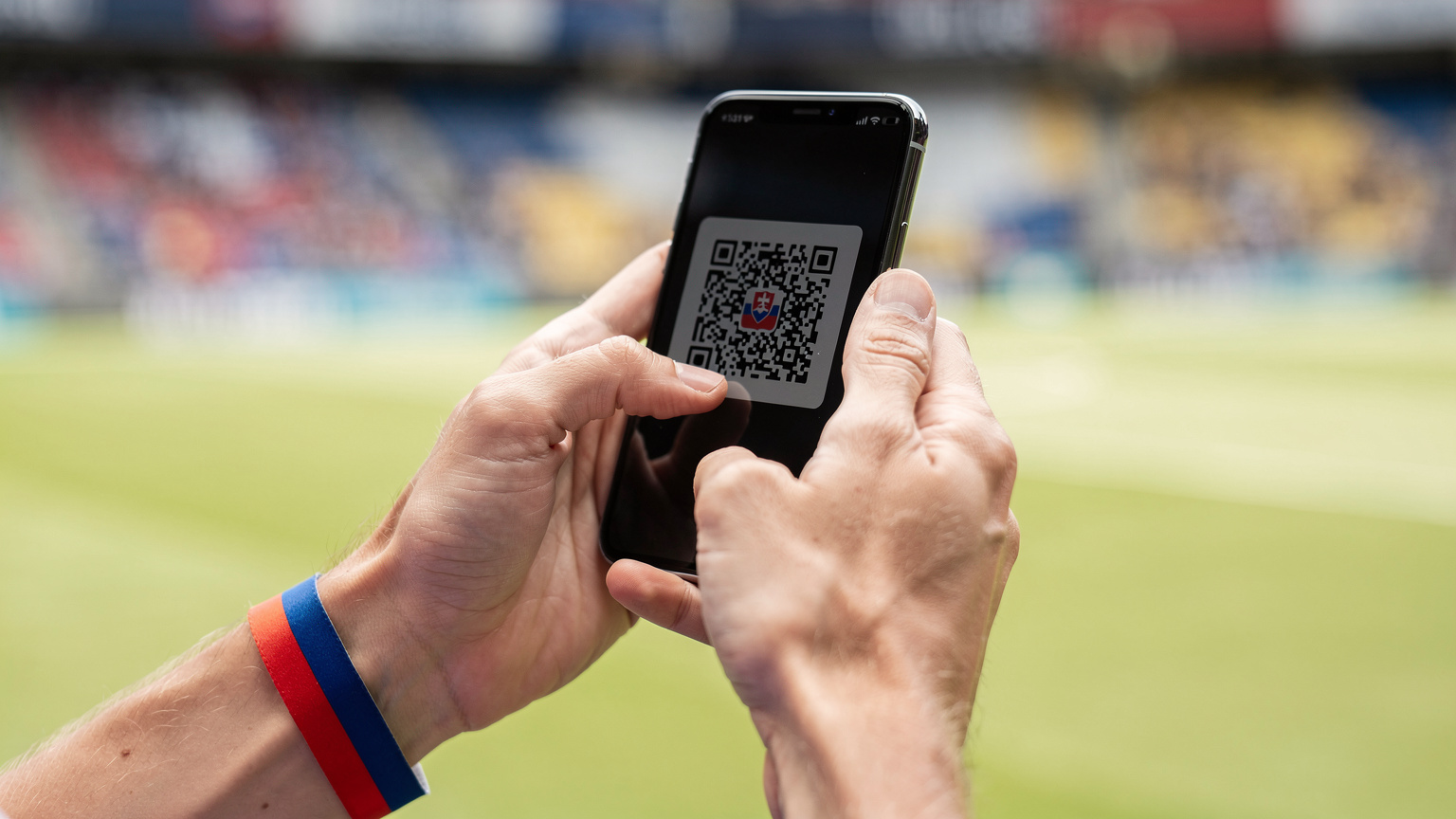 Detailný záber na ruky slovenského fanúšika, ktorý skenuje QR kód na aktiváciu eSIM na svojom smartfóne, s futbalovým štadiónom v pozadí.