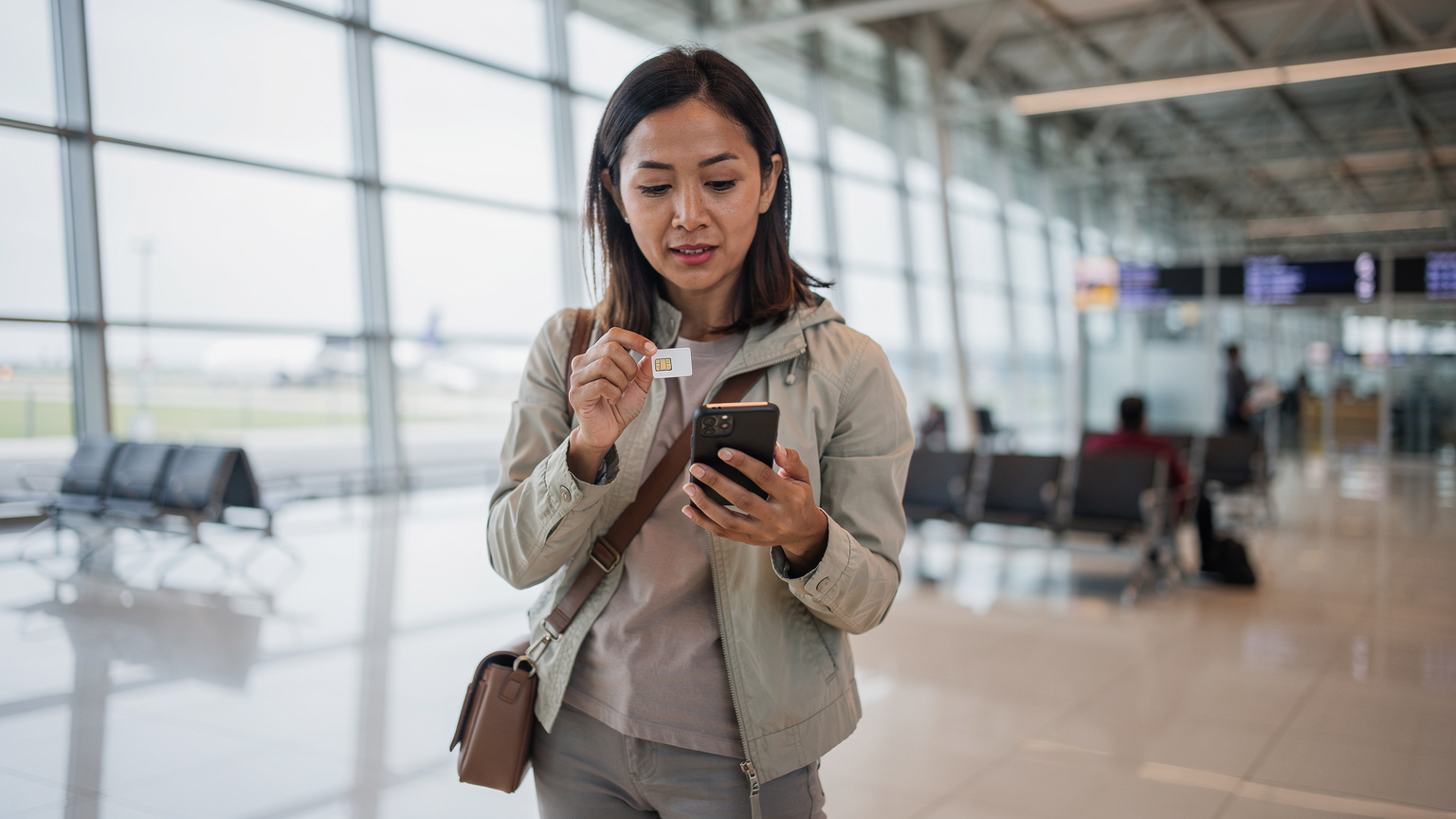 Seorang traveler sedang memegang ponselnya dan membandingkan eSIM dengan kartu SIM fisik di sebuah terminal bandara yang modern dan sepi.