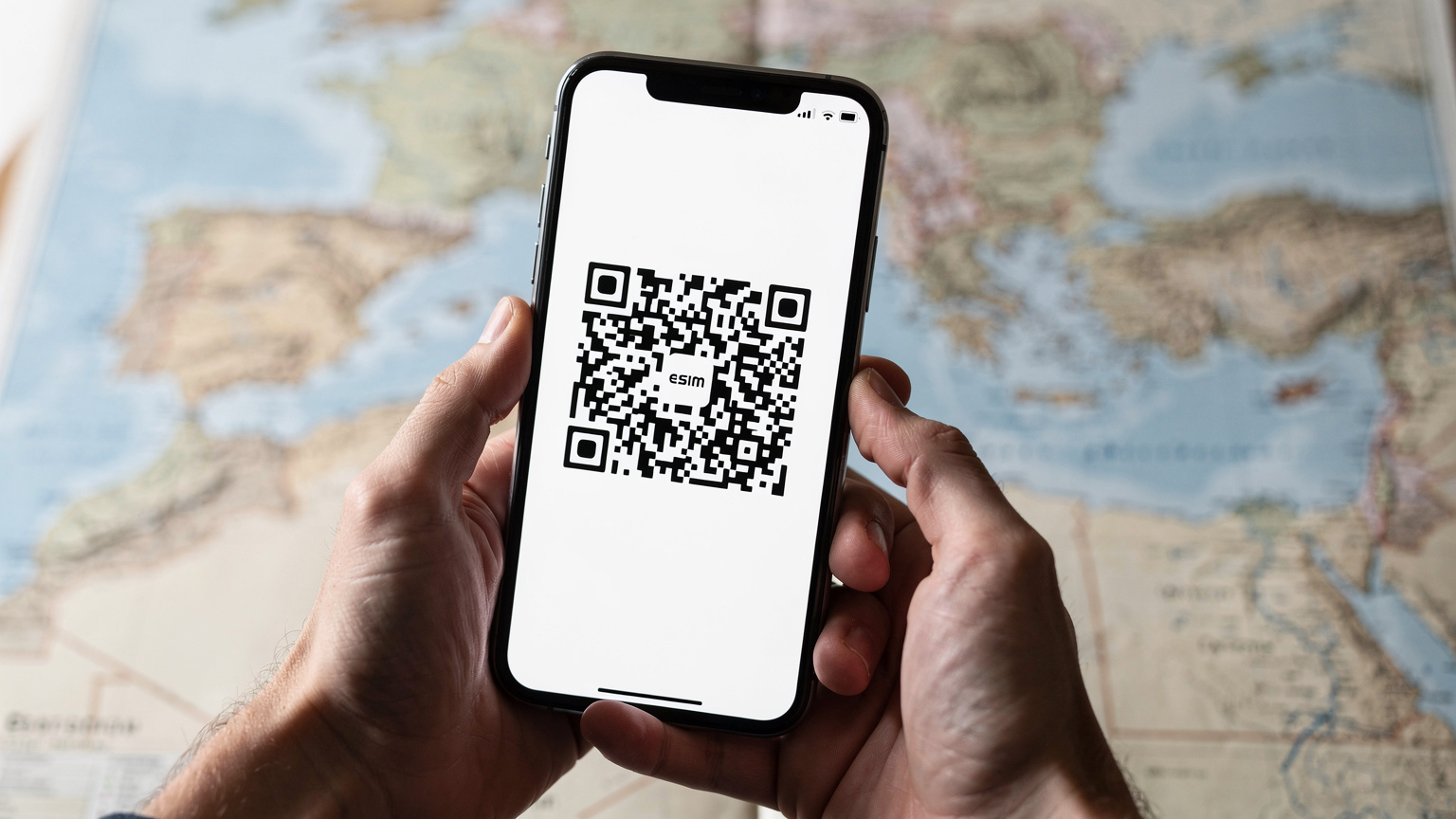 Un primo piano di mani che tengono uno smartphone moderno, mostrando l'interfaccia di attivazione di un profilo eSIM con un codice QR sullo schermo, mentre una mappa sfocata del Mediterraneo è visibile sullo sfondo.