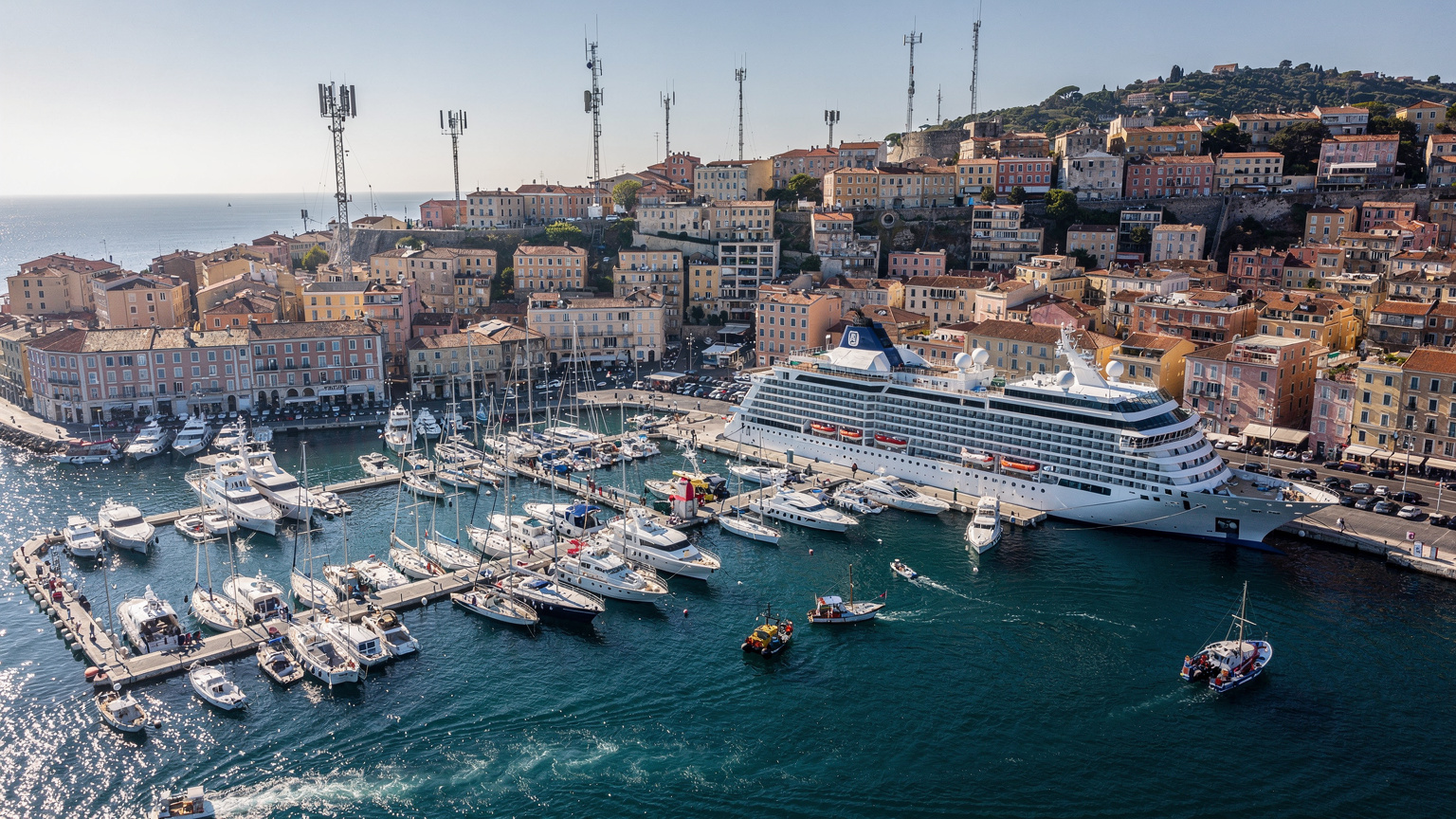 Una veduta aerea di un porto turistico affollato nel Mediterraneo, con yacht e navi da crociera ormeggiate, e un intreccio di antenne cellulari visibili sui tetti degli edifici circostanti, suggerendo una forte connettività di rete.