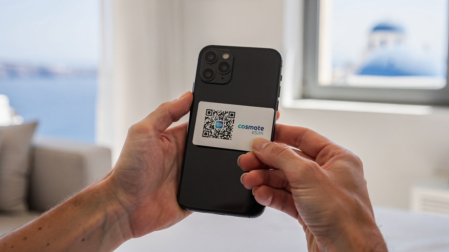 A close-up shot of hands holding a smartphone, with a Cosmote eSIM QR code on a piece of paper being scanned by the phone's camera against a backdrop of a hotel room in Santorini.