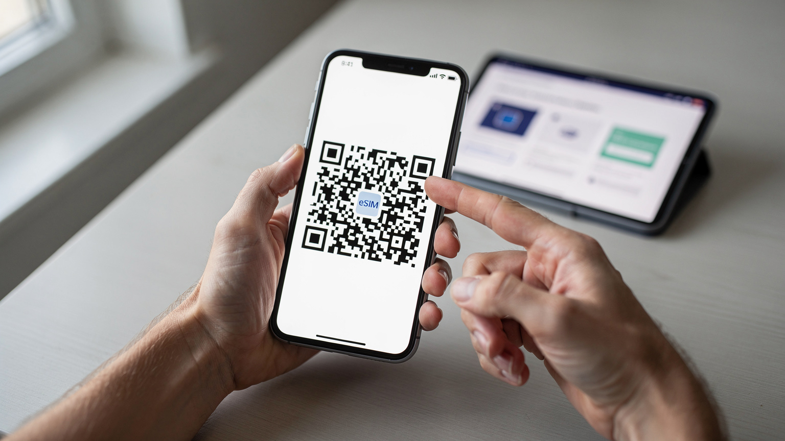 Primo piano sulle mani di una persona che tiene uno smartphone e mostra il codice QR per l'attivazione di una eSIM. Sullo sfondo, un tablet sfocato mostra i dettagli di un piano dati, evidenziando la semplicità del processo di attivazione digitale.