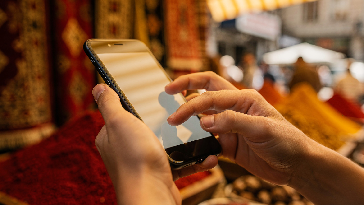 Close-up of hands interacting with a smartphone, tapping through mobile settings, with a blurry backdrop of a vibrant market stall in Istanbul, Turkey.