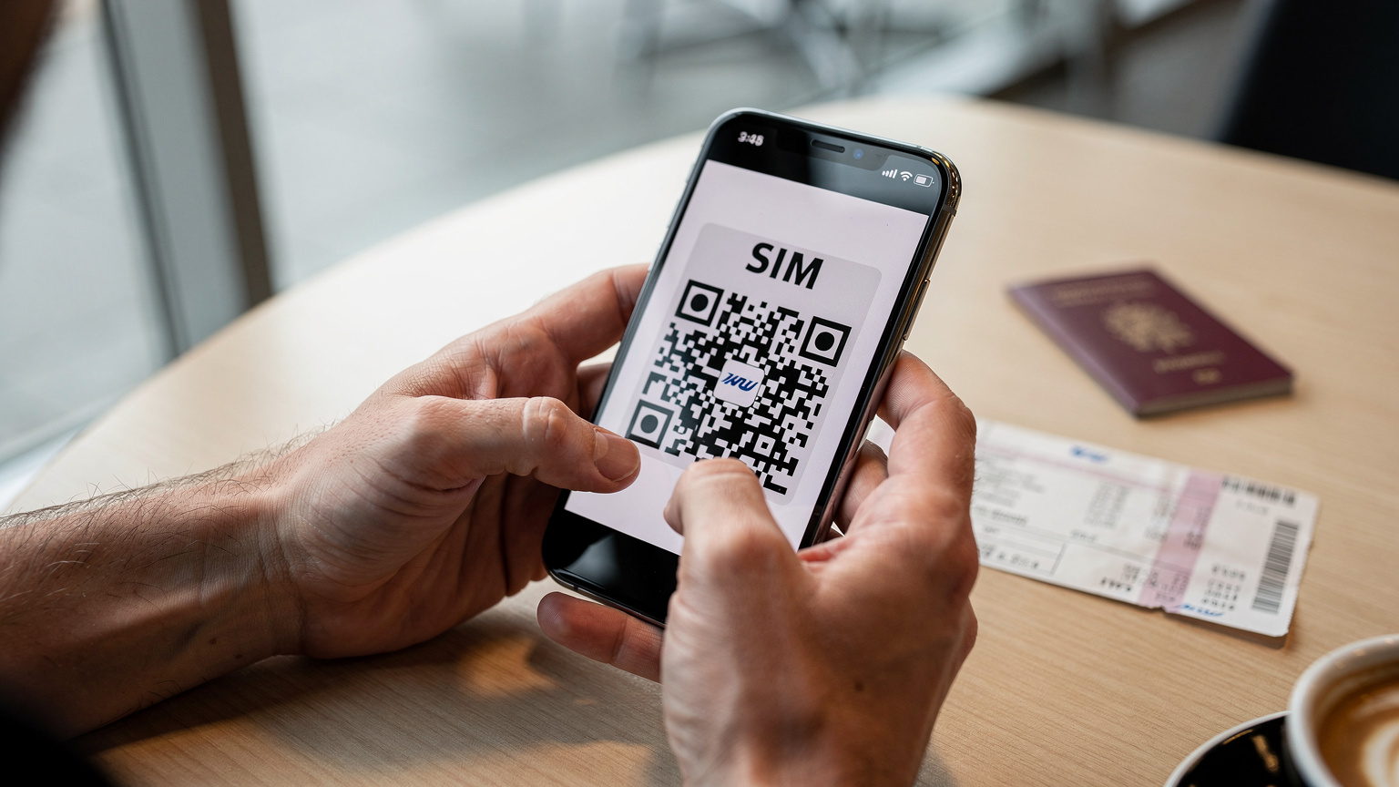Un gros plan sur des mains utilisant un smartphone pour scanner un QR code eSIM, avec un passeport français et un billet d'avion en arrière-plan flou, sur une table en bois clair d'un café d'aéroport. La lumière est douce et indirecte, venant d'une fenêtre.