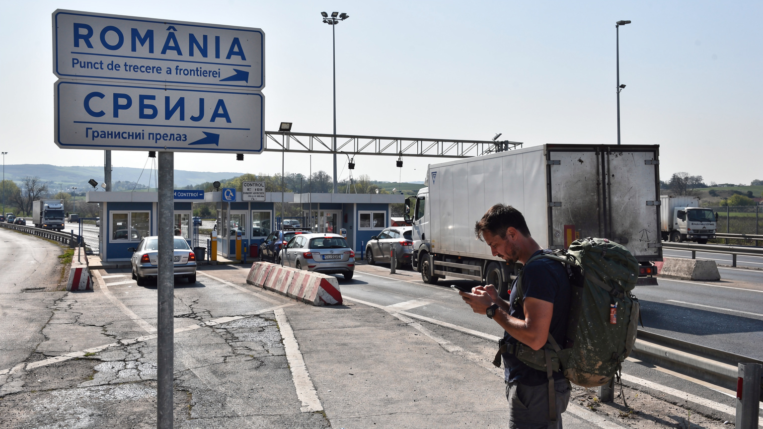 Un panou de semnalizare bilingv, română și sârbă, la un punct de trecere a frontierei, cu un călător verificând telefonul.