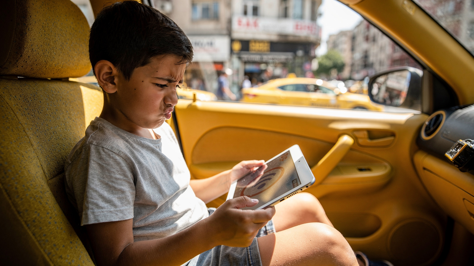Un băiețel de 7 ani se uită cu o expresie confuză la o tabletă care afișează o eroare de conexiune la internet, în timp ce stă pe bancheta din spate a unui taxi în aglomerația din Istanbul.