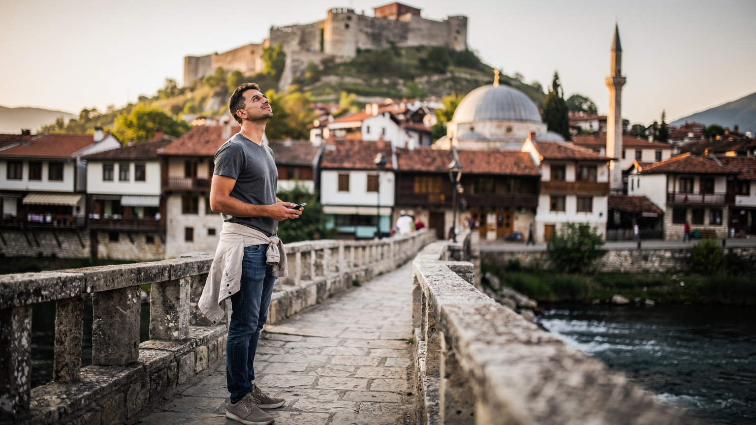 Prizren'in tarihi Şadırvan Meydanı'nda, Taş Köprü'nün üzerinde durmuş, elindeki telefonla kaleye doğru bakan genç bir adam.