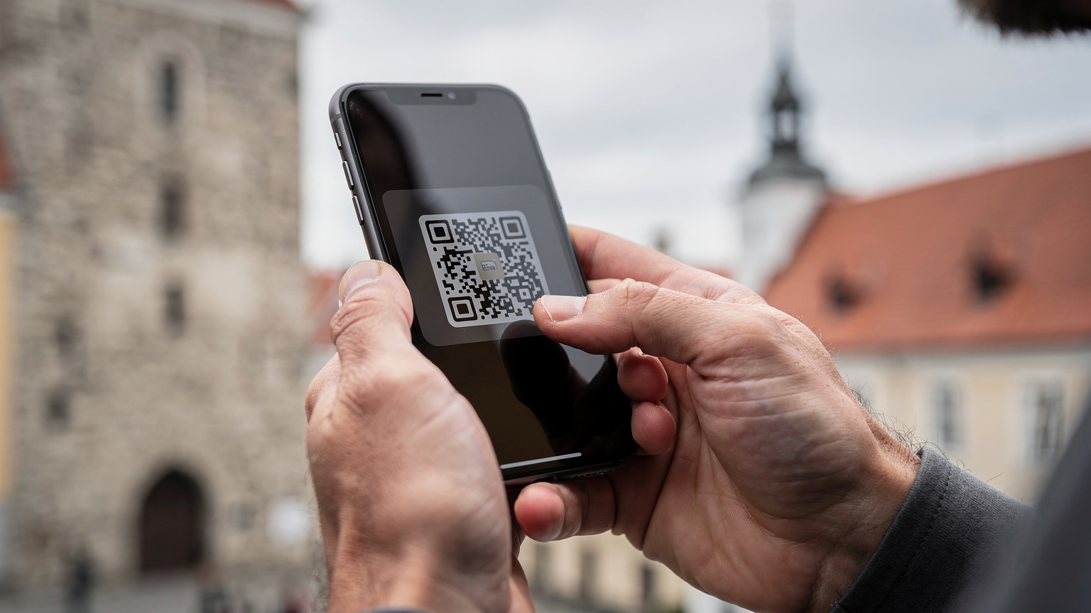 Крупный план рук, настраивающих eSIM на смартфоне, с размытым фоном старинных зданий Таллинна, Эстония.