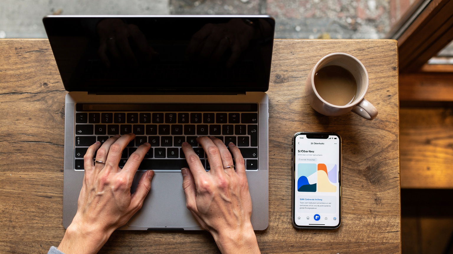 Nahaufnahme der Hände einer Person, die auf einem Laptop im Co-Working Space 'St. Oberholz' in Berlin tippt, während ihr Smartphone mit einer eSIM-Anbieter-App daneben liegt und eine Kaffeetasse in Reichweite ist.