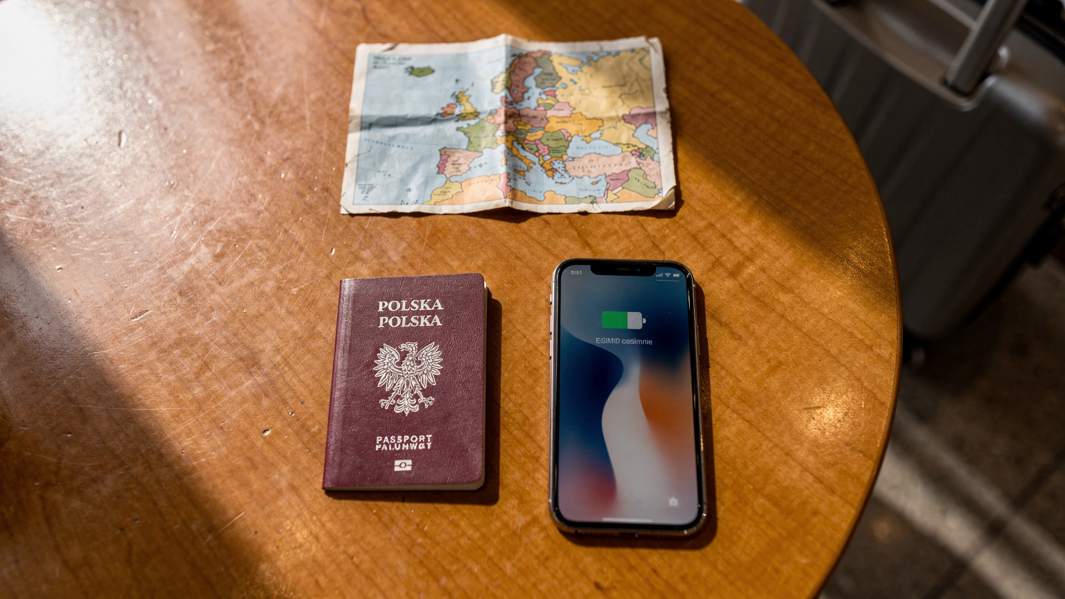 Paszport, smartfon z aktywną aplikacją eSIM i mała mapa Europy leżą na drewnianym stoliku w kawiarni na lotnisku, z walizką w tle.