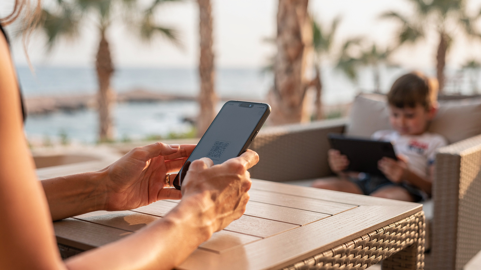 O mamă, cu un copil mic care se joacă liniștit pe o tabletă lângă ea, scannând un cod QR cu telefonul său pentru a activa un eSIM, pe o terasă relaxantă a unui hotel din Sharm el-Sheikh.