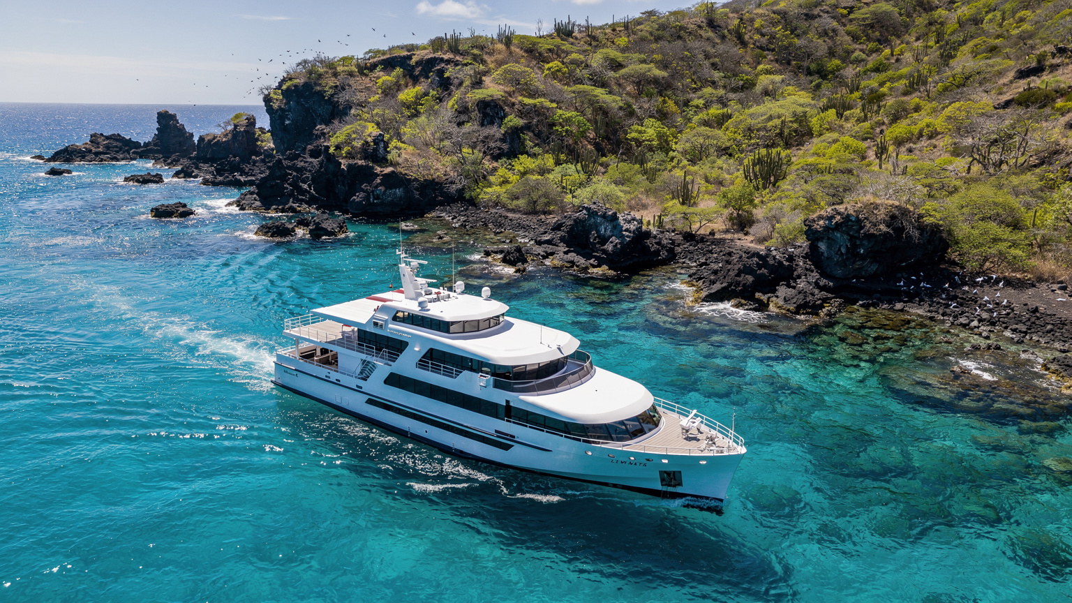 Ein modernes Kreuzfahrtschiff gleitet durch ruhiges, türkisfarbenes Wasser in der Nähe der Galápagos-Inseln, umgeben von üppiger, unberührter Natur.