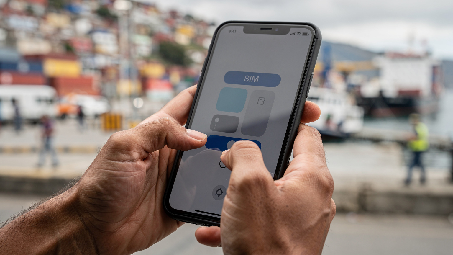 Eine Nahaufnahme zeigt Hände, die ein Smartphone halten, auf dem eine eSIM-Aktivierungsseite angezeigt wird, während im Hintergrund ein Hafen in Südamerika unscharf zu sehen ist.