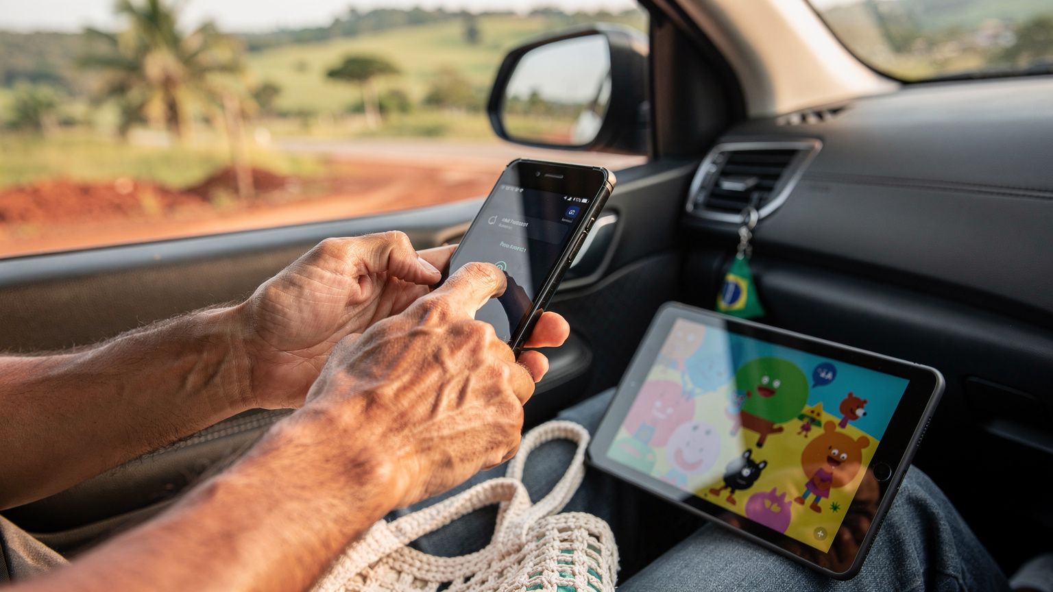 Uma mão adulta configurando um smartphone para compartilhar dados via hotspot, com um tablet infantil ao lado exibindo um desenho animado, no interior de um carro em viagem pelo Brasil.