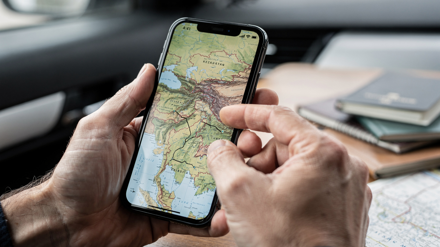 A close-up shot of a traveler's hands holding a smartphone, with a detailed map of Central Asia displayed on the screen, showing various country borders and a planned multi-country route.