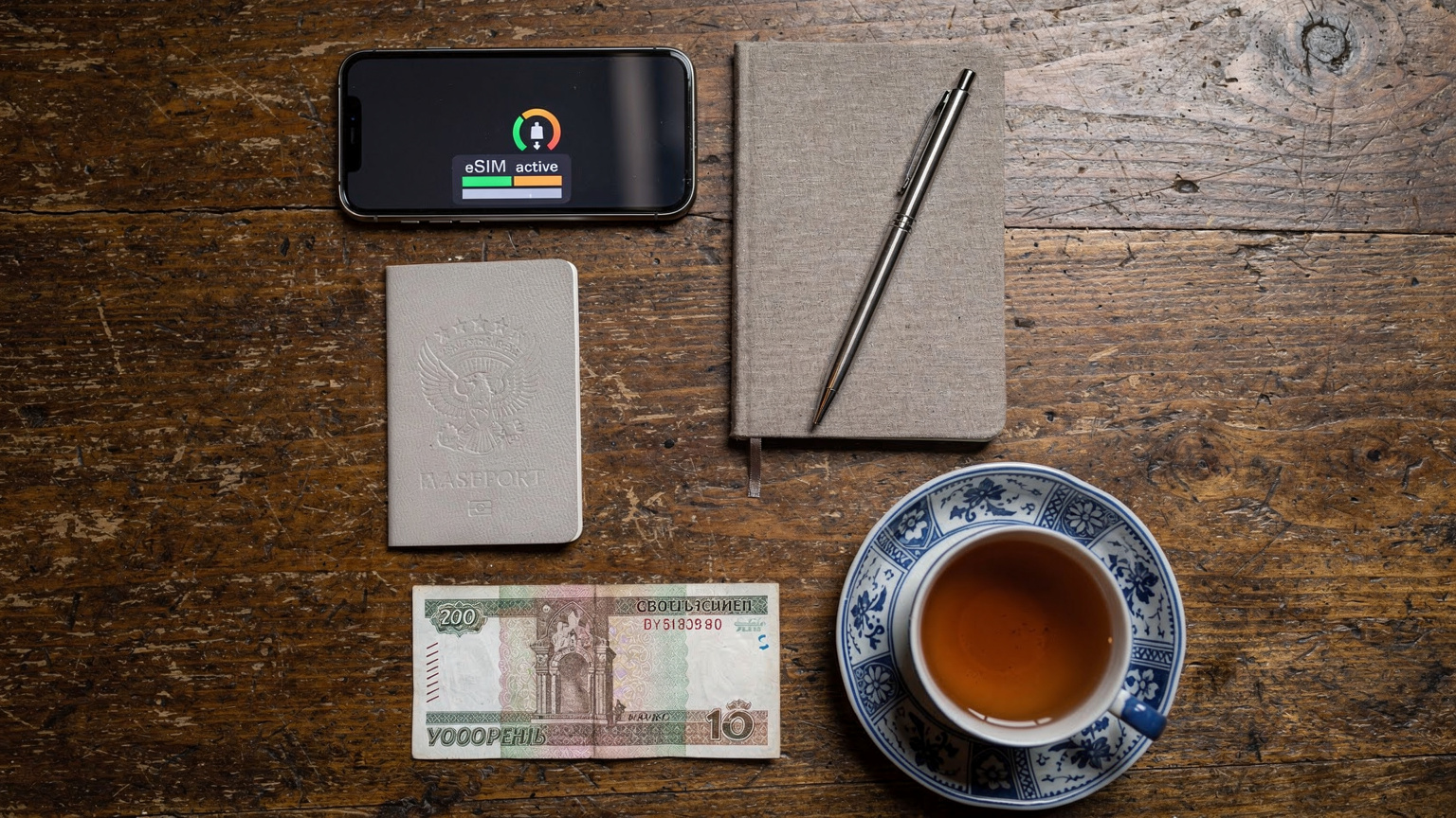 An overhead flat-lay of travel essentials for Central Asia: a passport, a smartphone showing an active eSIM profile, a small notebook, a local currency banknote (Uzbek Sum or Kazakh Tenge), and a cup of traditional Central Asian tea on a wooden table.