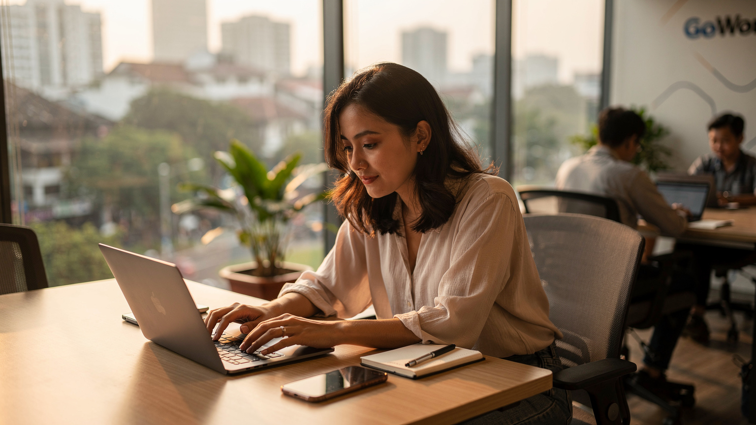 Seorang pengembara sedang menaip di komputer ribanya di sebuah co-working space yang moden di Jakarta, dengan telefon pintarnya diletakkan di sebelahnya, menunjukkan kepentingan sambungan internet yang stabil.