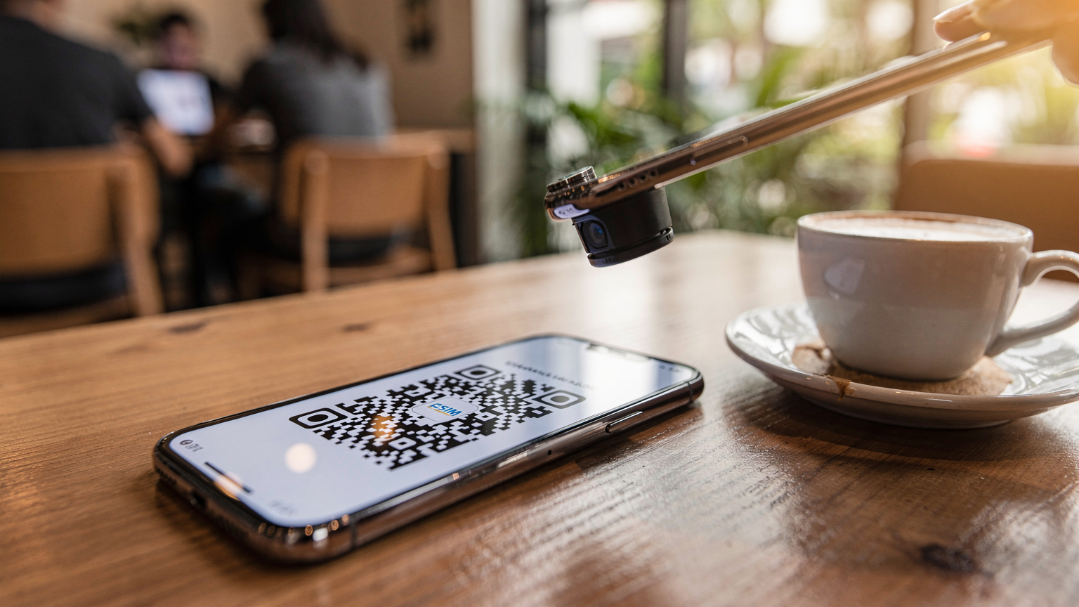 Sebuah ponsel yang menampilkan QR code di layarnya sedang dipindai oleh ponsel lain, dengan latar belakang meja kopi di sebuah kafe bergaya.
