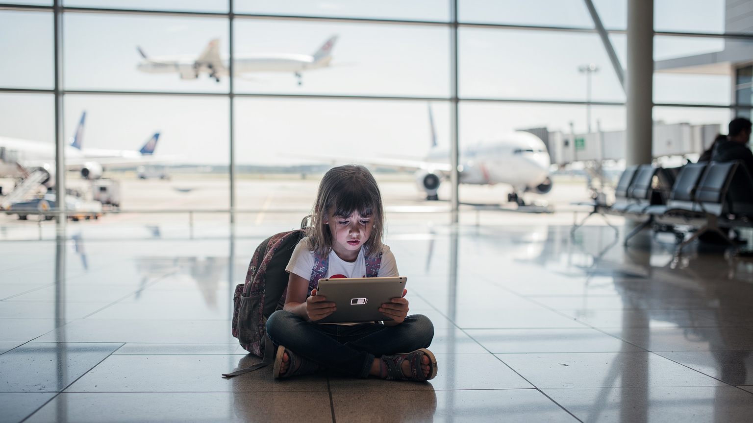Ein kleines Mädchen sitzt auf dem Boden vor einem großen Flughafenfenster und schaut sorgenvoll auf ihr Tablet, dessen Bildschirm ein leeres Akku-Symbol zeigt, während im Hintergrund Flugzeuge starten und landen.