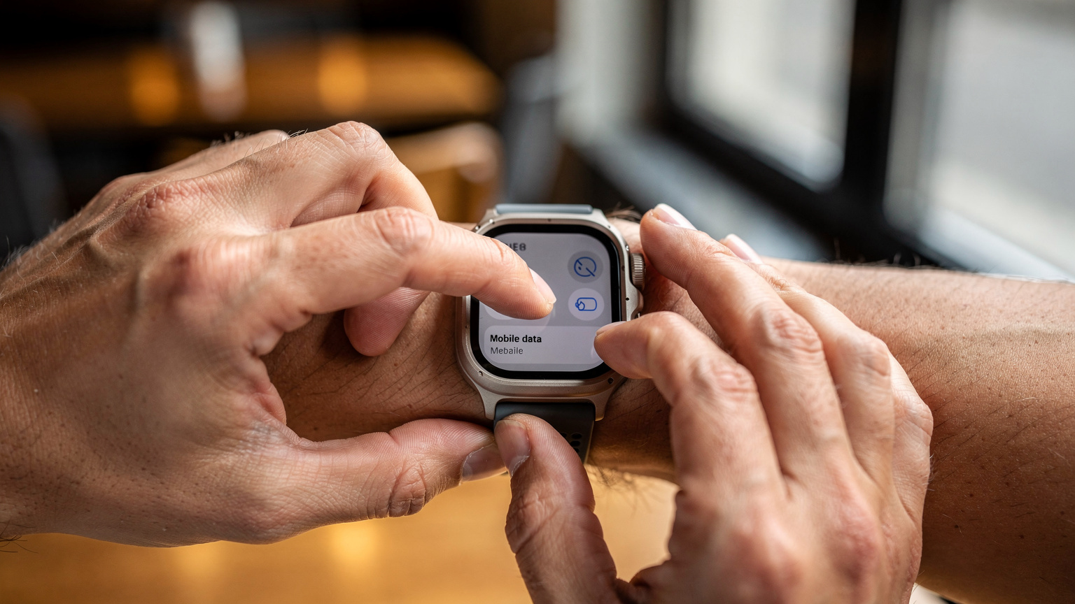 Nahaufnahme der Hände einer Person, die auf der Apple Watch Ultra 3 durch die Einstellungen scrollt, um mobile Daten zu deaktivieren oder zu konfigurieren, mit einem verschwommenen Hintergrund einer Café-Umgebung.