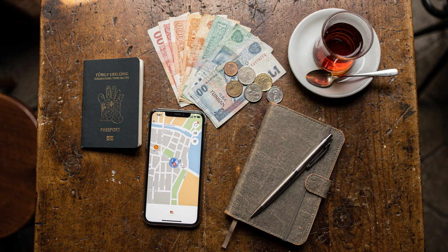 An overhead flat-lay of travel essentials: passport, smartphone with a digital map, local currency, and a small notebook, on a café table in Istanbul.