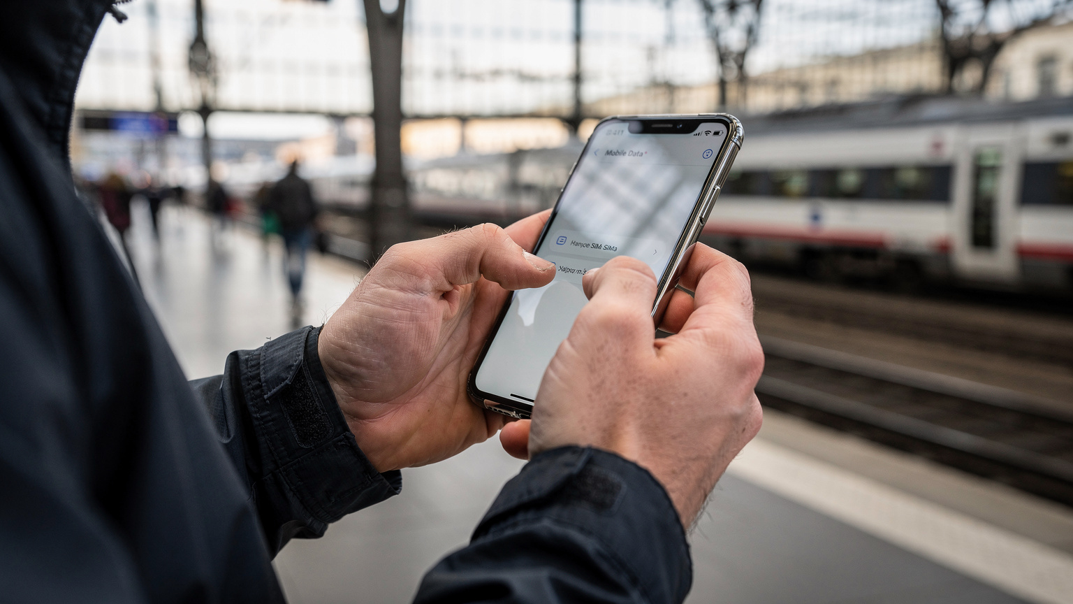 Close-up of hands adjusting dual-SIM settings on a smartphone, with a blurred backdrop of a train station platform.