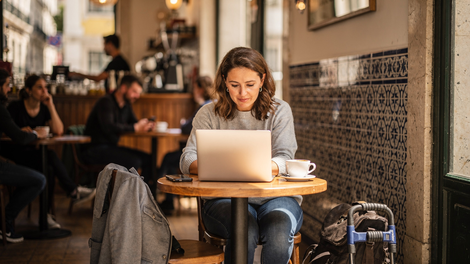 Uma mulher em um café movimentado em Lisboa, Portugal, usando seu laptop e smartphone, com um cadeado de bicicleta preso à sua mochila, simbolizando a atenção à segurança digital e física durante a viagem.