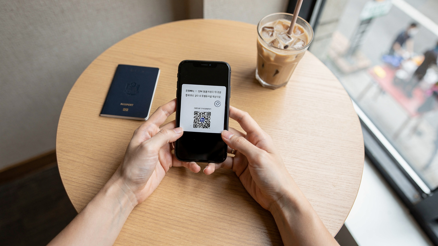 韓国のカフェで、旅行者がスマートフォンを使ってCellesimのeSIM設定をQRコードで読み取っている様子。手元にはパスポートとコーヒーが置かれ、リラックスした雰囲気。