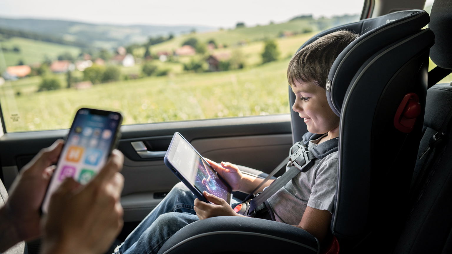 Un copil fericit se joacă pe o tabletă într-o mașină, cu peisajul rural bulgăresc în fundal, iar părintele din față monitorizează discret activitatea pe telefonul său.
