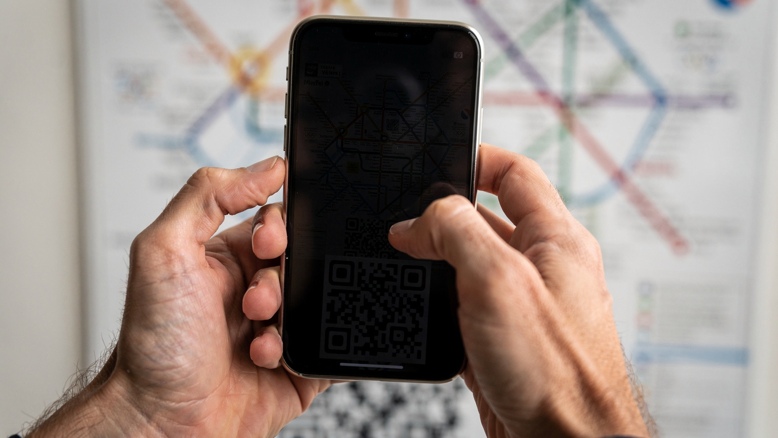 Gros plan sur des mains scannant un QR code d'eSIM avec un iPhone, avec en arrière-plan flou une carte du métro parisien et le logo des JO 2026.