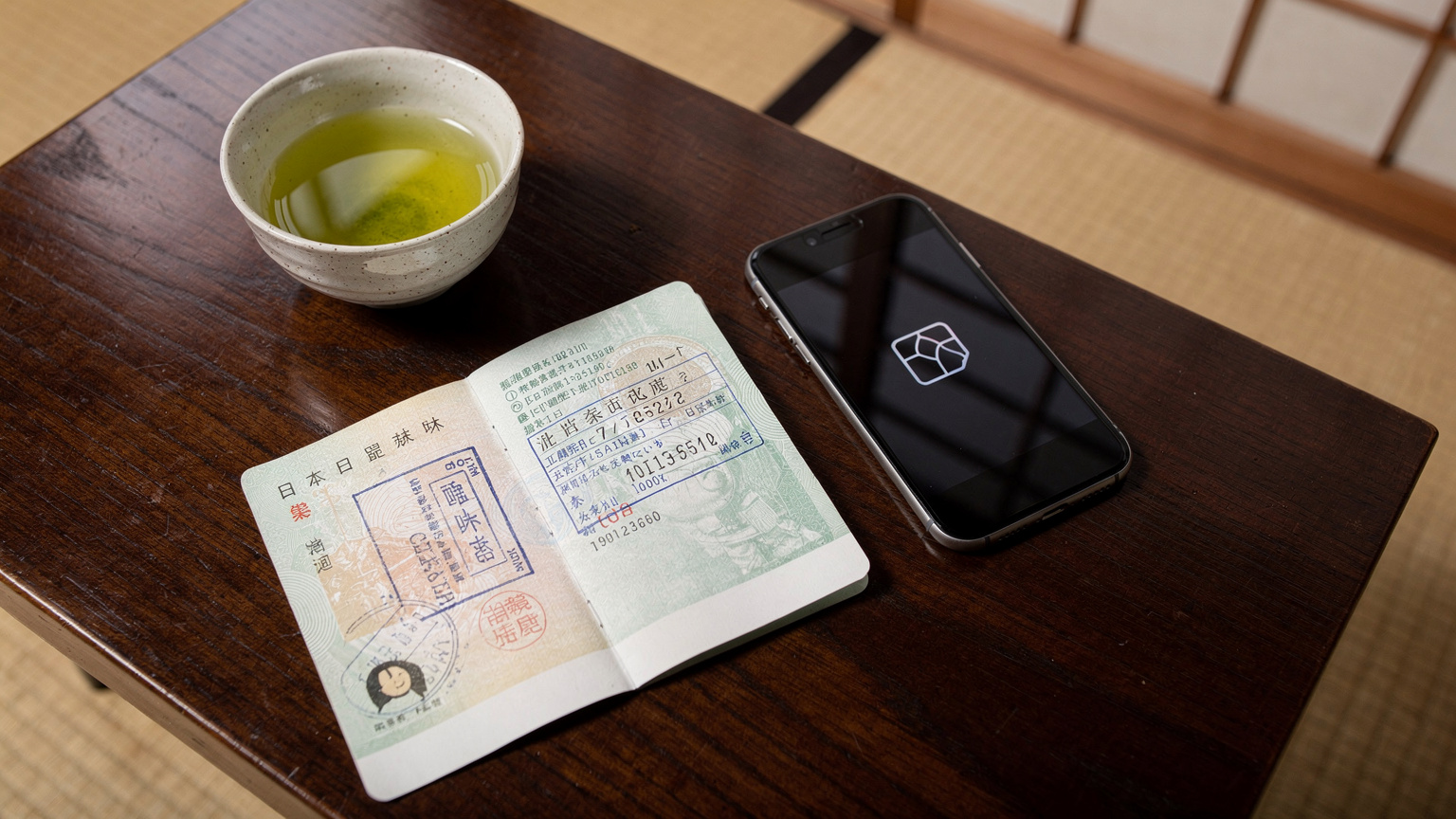 Un passaporto italiano aperto su una pagina del visto giapponese, appoggiato su un tavolo di legno in un tradizionale ryokan, con un moderno smartphone e una tazza di tè verde accanto.