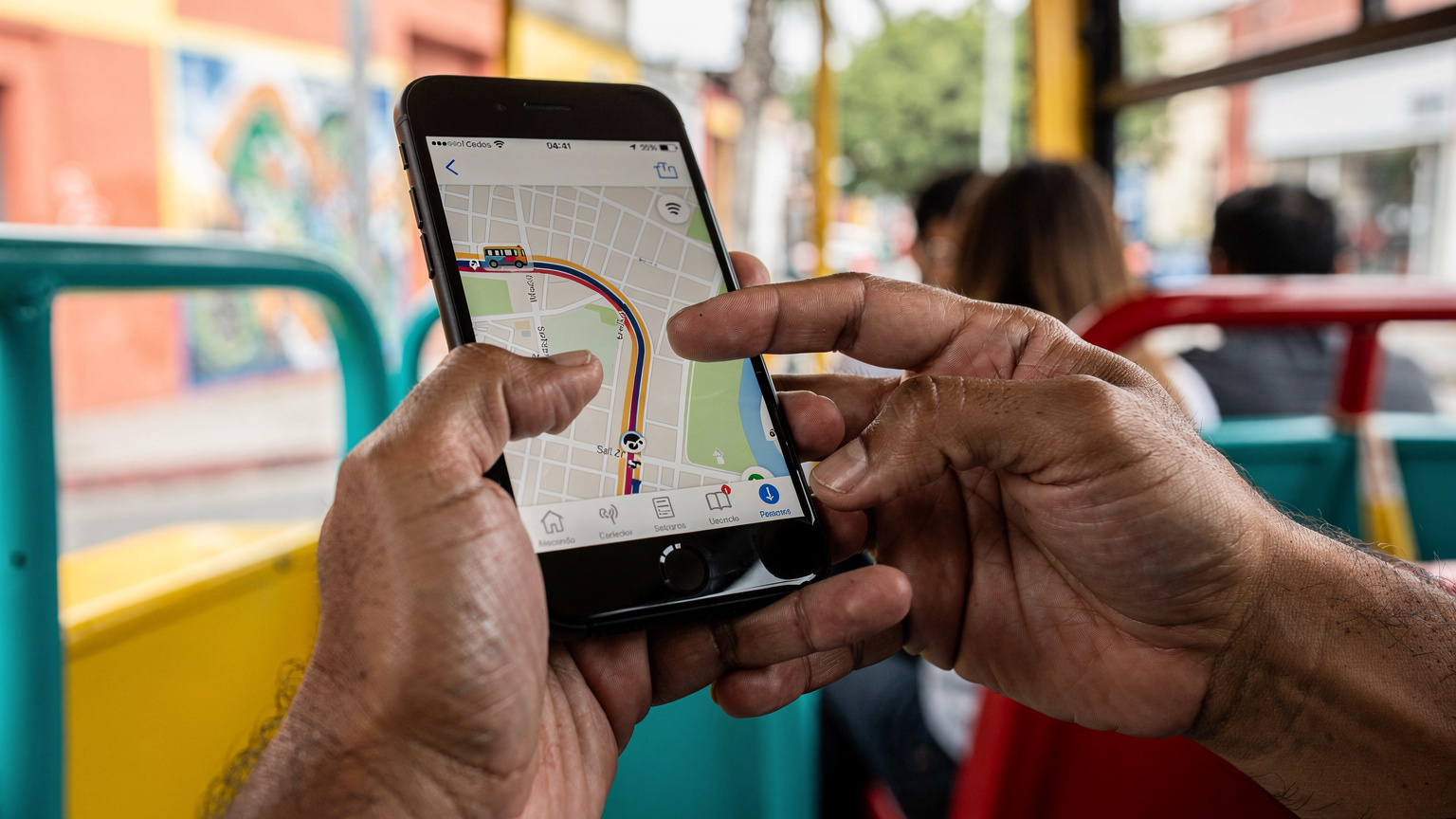 Gros plan sur des mains utilisant une application de covoiturage sur un smartphone, l'interface affichant un itinéraire de bus sur une carte mexicaine, dans un bus de transport public coloré et légèrement bondé.