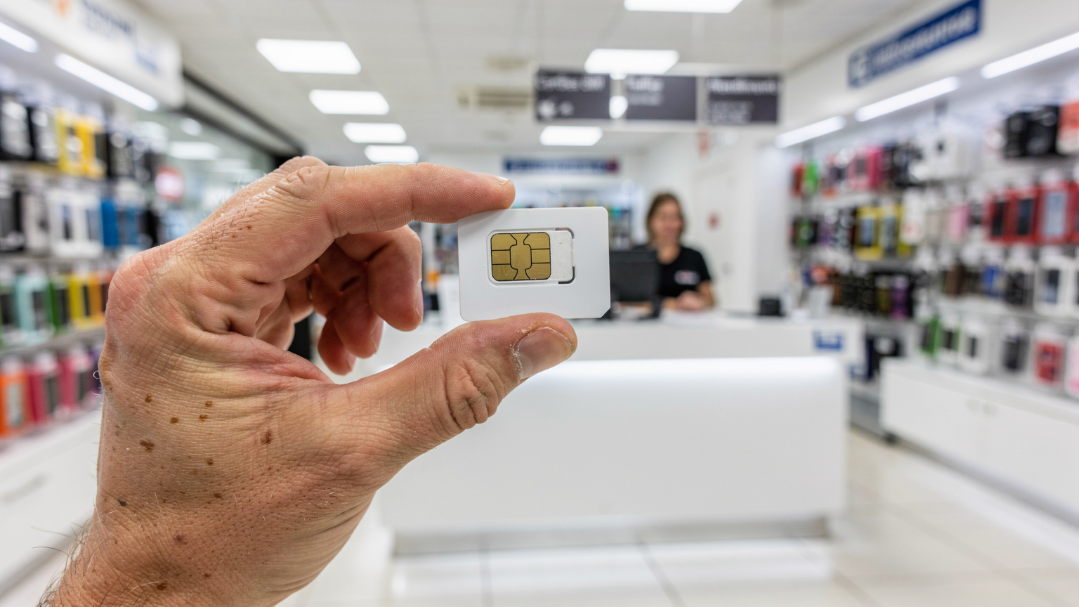 Uma mão segurando um cartão SIM físico português recém-comprado, com o balcão de atendimento de uma loja de operadora ao fundo.