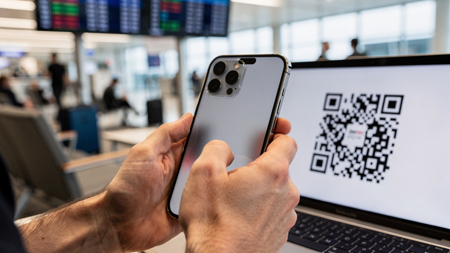 A close-up of a traveller's hands holding an iPhone, scanning an eSIM QR code from a laptop screen in a bustling airport lounge.