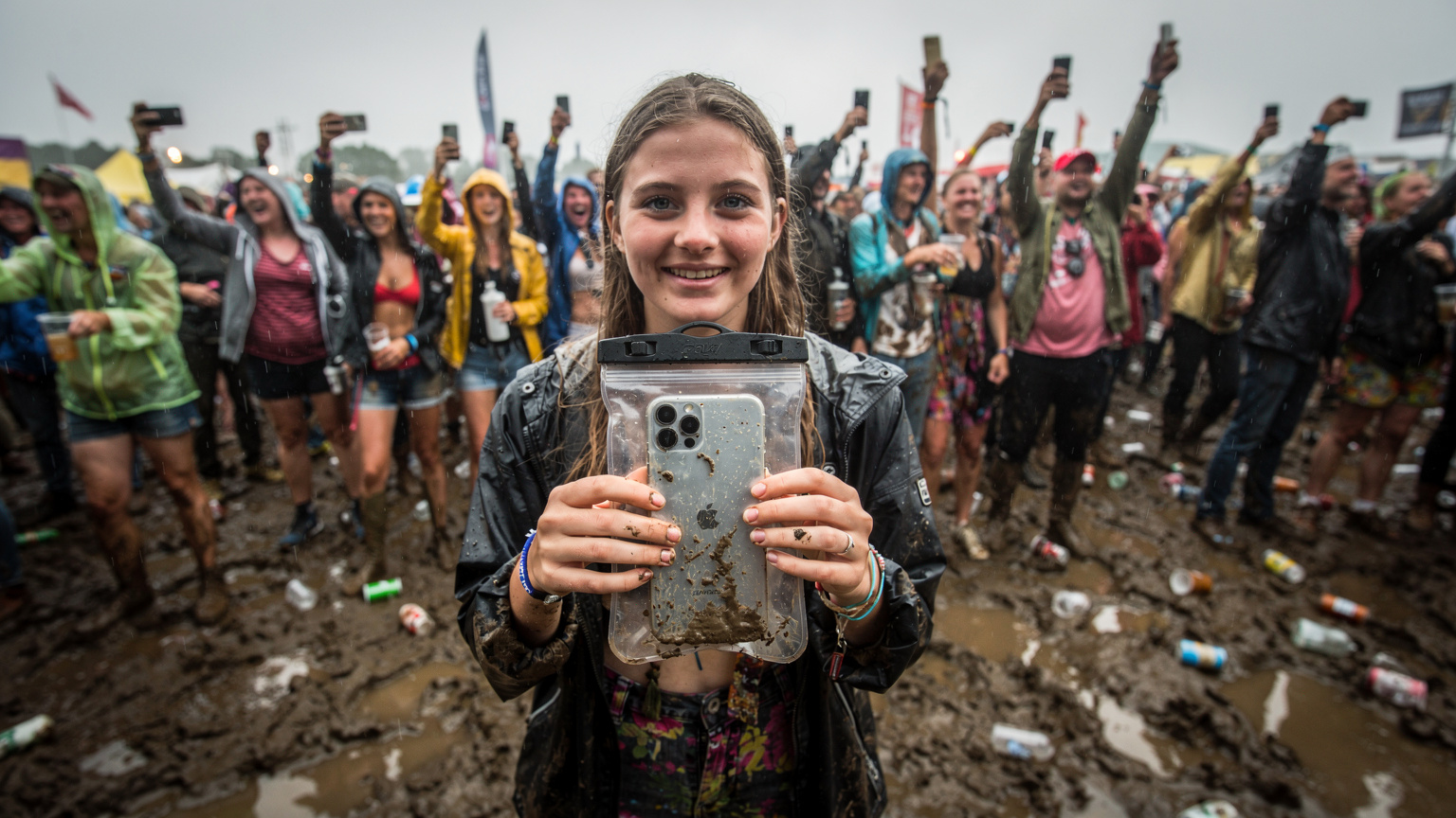 Eine junge Frau schützt ihr Smartphone, das in einer transparenten, wasserdichten Hülle steckt, während sie auf einem schlammigen Festivalgelände zwischen tanzenden Menschen steht, die ihre eigenen Smartphones in die Luft halten.
