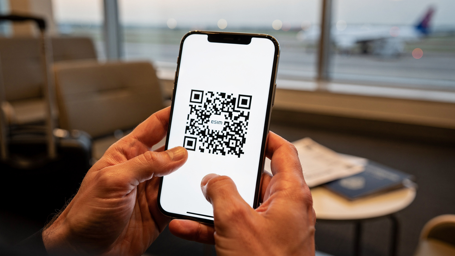 Manos de un viajero sosteniendo un smartphone, mostrando la pantalla con un código QR de una eSIM listo para ser escaneado, en un entorno de aeropuerto.