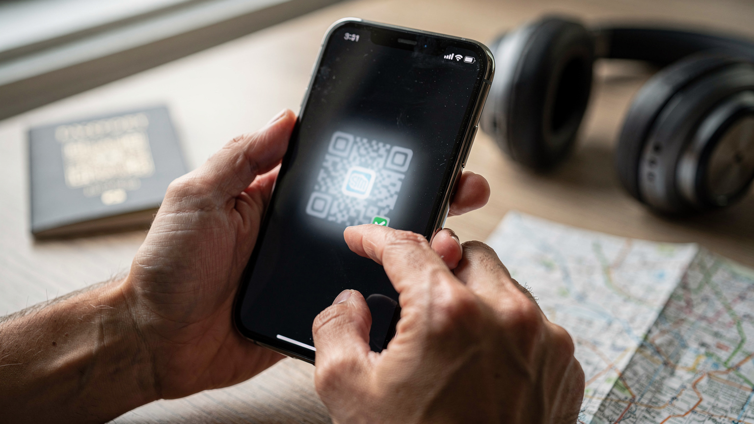 Manos de una persona interactuando con una pantalla de teléfono, mostrando el proceso de escaneo de un código QR para activar una eSIM. Las manos son de un color de piel cálido, y el teléfono es moderno. El fondo es un escritorio borroso con elementos de viaje como un pasaporte o un mapa.